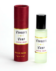 A small Cowboy's Camp Perfume Oil bottle by Faire, filled with pale essential oils and topped with a black cap, stands beside its maroon and gold cylindrical box.
