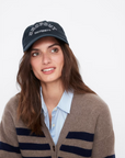 A person with long brown hair is wearing a black, 100% cotton baseball cap titled "THE DROPOUT KAP NAVY" by Kule. They are dressed in a light blue collared shirt and a brown cardigan with blue stripes, all set against a plain white background.