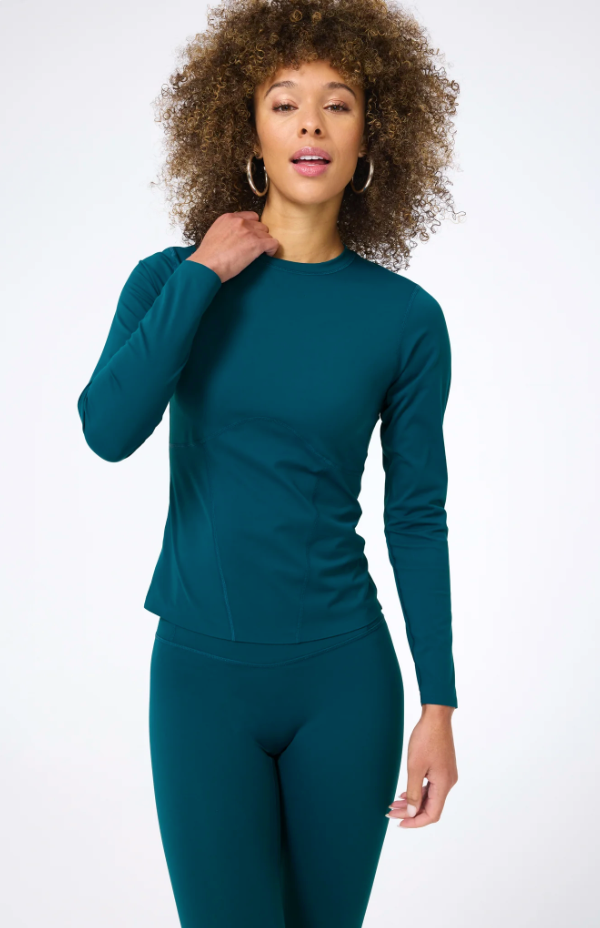 A woman with curly hair wears a Terez Action Corset Long Sleeve Top in Cypress and leggings, posing against the white walls of a Scottsdale, Arizona bungalow.