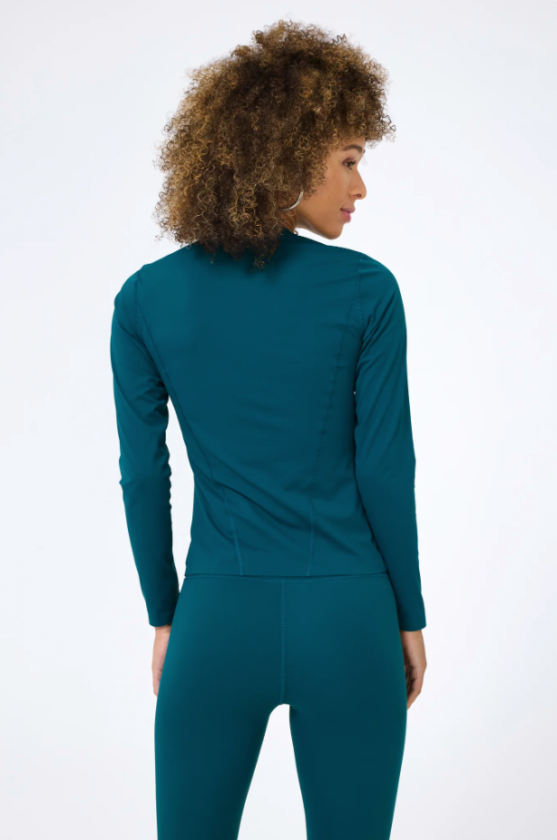 A woman with curly hair seen from the back wearing a teal turtleneck top and matching pants stands against a solid white background in a Terez Action Corset Long Sleeve Top in Cypress.
