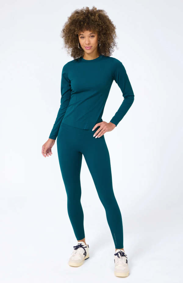 A woman with curly hair stands confidently in Scottsdale, Arizona, wearing a Terez Action Corset Long Sleeve Top in Cypress and leggings, paired with white sneakers against a white background.