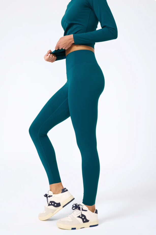A woman in a teal long-sleeve top and Terez's Action Basic Legging in Cypress, adjusting a fitness tracker on her wrist, stands against a plain white background. Only her torso and legs are visible, reminiscent of the relaxed vibe found in Scottsdale, Arizona.
