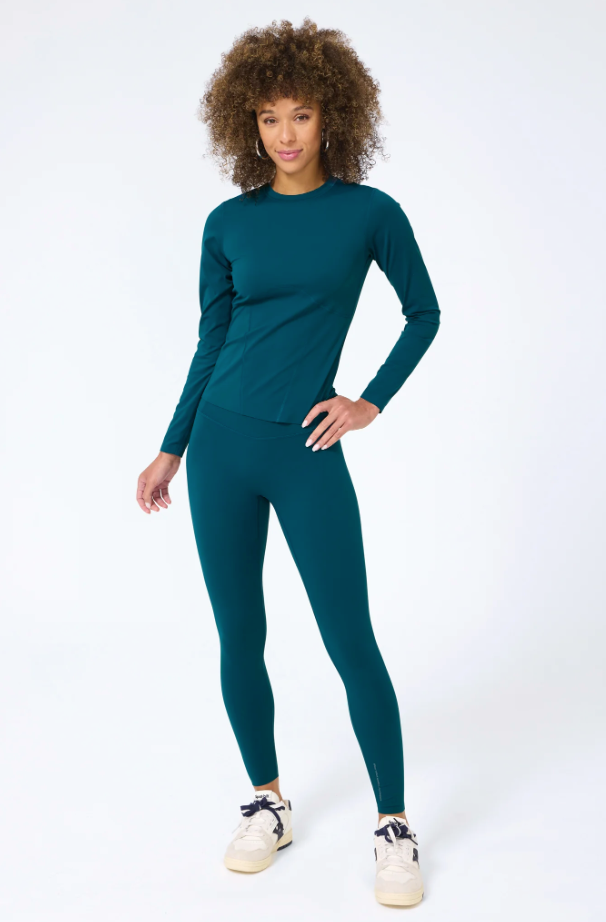 A woman with curly hair wearing a teal activewear outfit, including a long-sleeve top and Terez Action Basic Legging in Cypress, standing confidently with one hand on her hip against the plain white background of a Scottsdale, Arizona bungalow.