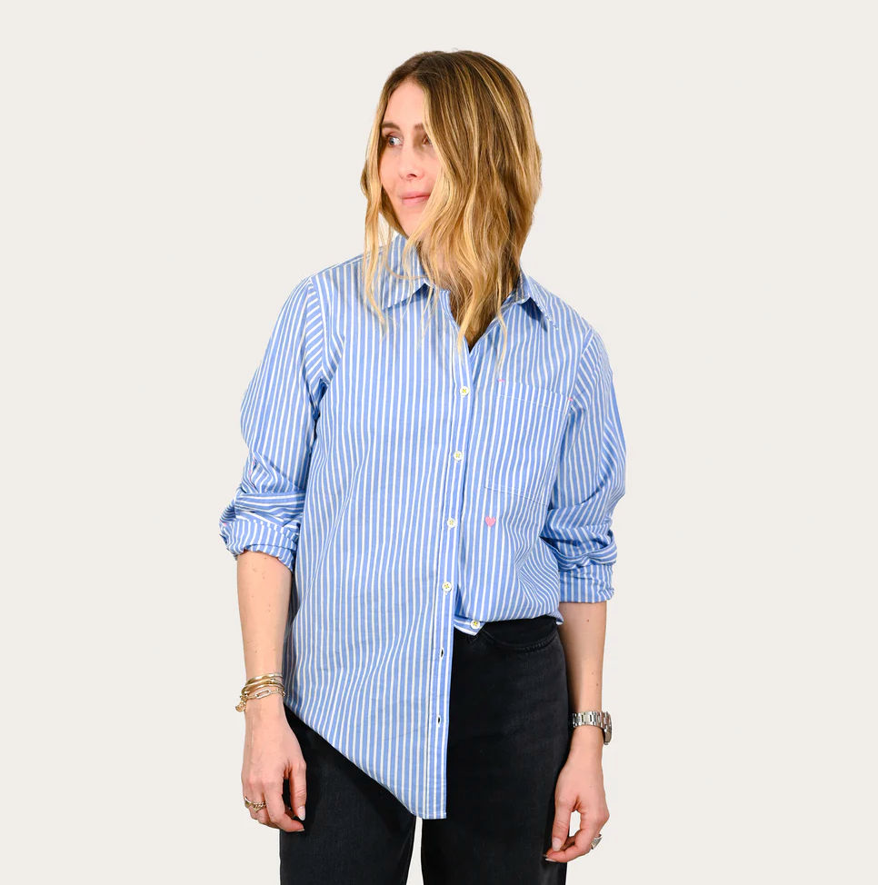 A woman with shoulder-length blonde hair stands against a white background, wearing a Mia Shirt Stripe Blue/white Blue by Kerri Rosenthal and black trousers, looking to her left with a slight smile.
