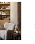 A cozy reading nook in a Scottsdale Arizona bungalow with a plush, light gray chair next to a wooden side table with a vase of dried flowers. A bookshelf filled with various decorative items is seen in the background, featuring "Call It Home: The Details That Matter" by Random House.