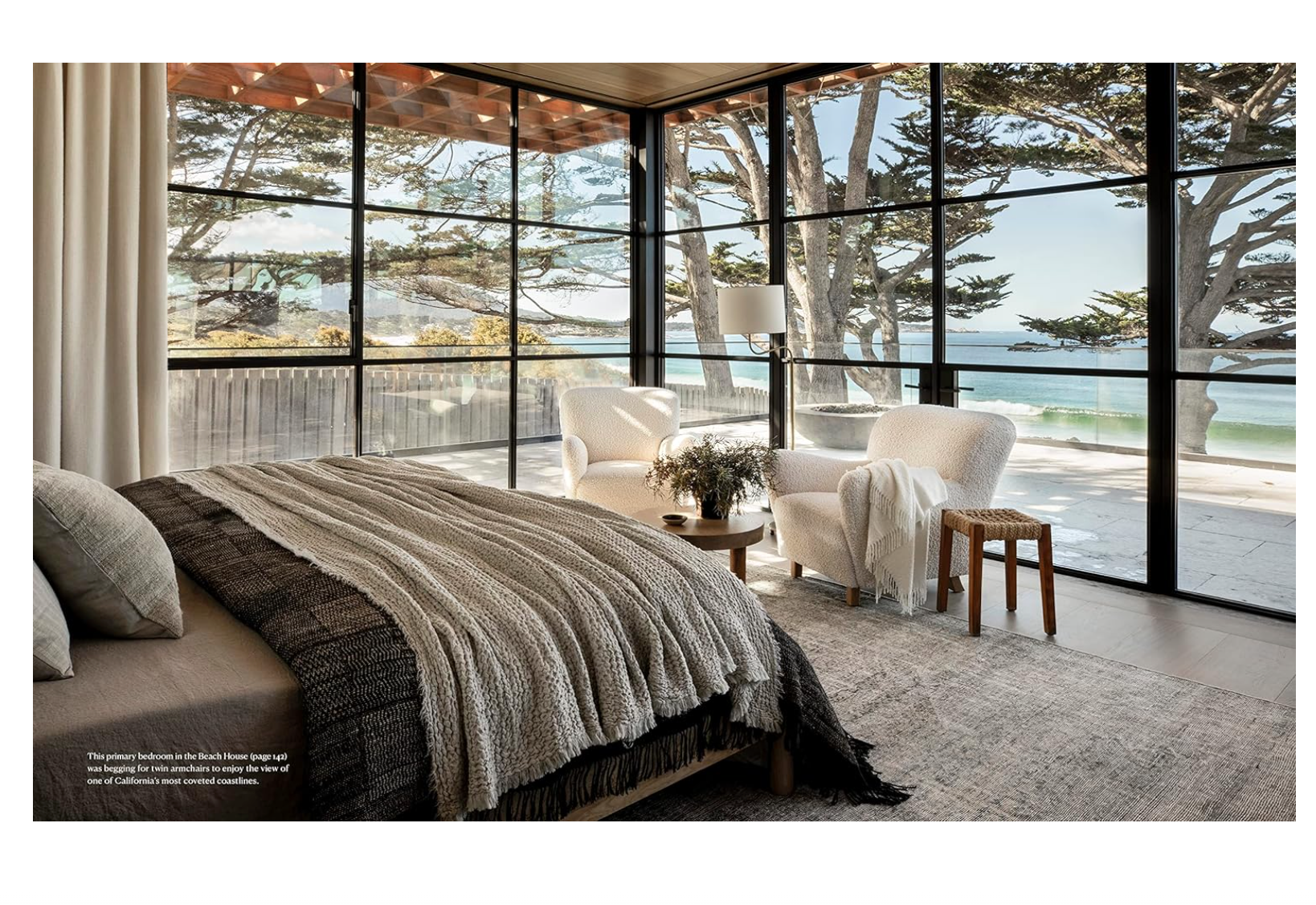 A serene bedroom in a bungalow with large windows overlooking a beach, featuring a cozy bed with a gray blanket, a plush armchair, and a small table with a vase and books. A floor lamp stands in the corner, all from Random House's "Call It Home: The Details That Matter.