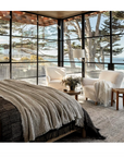 A serene bedroom in a bungalow with large windows overlooking a beach, featuring a cozy bed with a gray blanket, a plush armchair, and a small table with a vase and books. A floor lamp stands in the corner, all from Random House's "Call It Home: The Details That Matter.