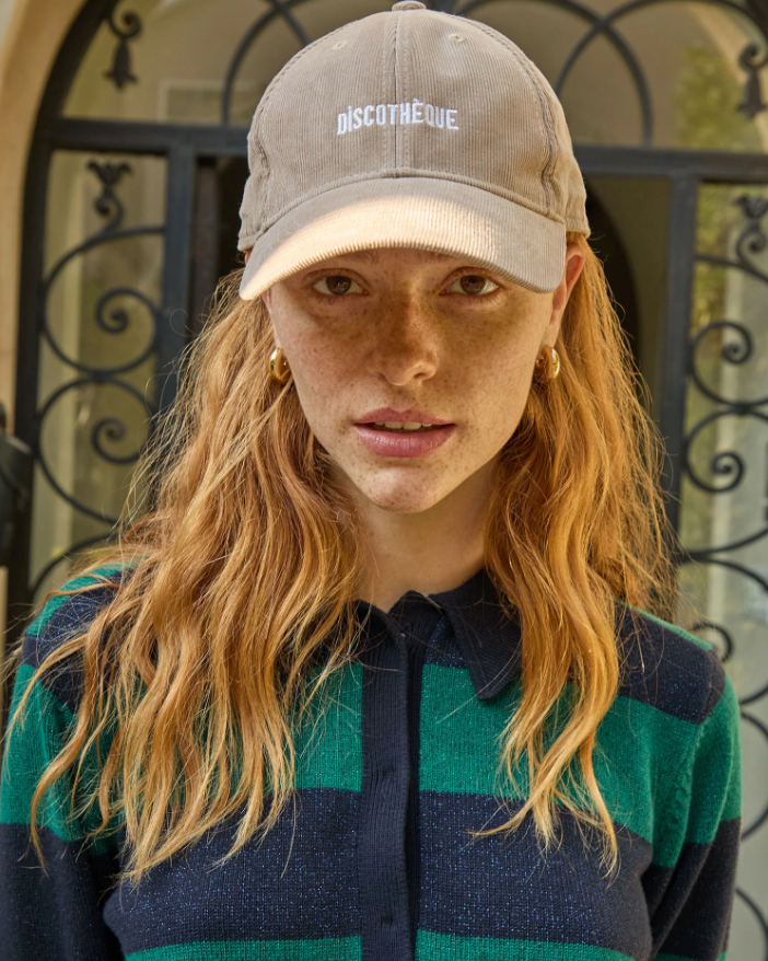 A woman with wavy red hair and freckles wearing a Clare Vivier Baseball Hat Stone Corduroy Discothèque and a green and black striped top, standing in front of a door with intricate designs in Arizona.