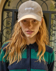 A woman with wavy red hair and freckles wearing a Clare Vivier Baseball Hat Stone Corduroy Discothèque and a green and black striped top, standing in front of a door with intricate designs in Arizona.