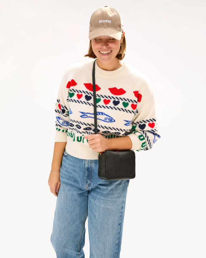 A smiling woman in a white sweater with colorful eye and lip patterns, blue jeans, a Baseball Hat Stone Corduroy Discothèque by Clare Vivier, and a black crossbody bag, embodying Arizona style, standing against a plain background.