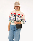 A smiling woman in a white sweater with colorful eye and lip patterns, blue jeans, a Baseball Hat Stone Corduroy Discothèque by Clare Vivier, and a black crossbody bag, embodying Arizona style, standing against a plain background.