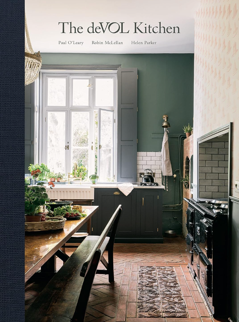 A cozy kitchen with dark green cabinets, white subway tile backsplash, and a vintage black stove. A large window bathes the space in natural light. Wooden counters adorned with fresh vegetables and potted plants showcase The deVOL Kitchen: Designing and Styling the Most Important Room in Your Home by Random House for any kitchen company enthusiast.