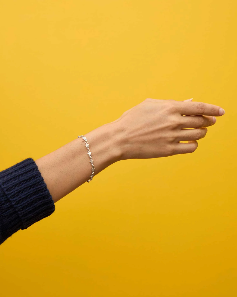 A woman&#39;s hand extended against a yellow background, featuring a Clare Vivier Star Strand Bracelet Sterling Silver on the wrist. She wears a navy blue sweater in true Arizona style.