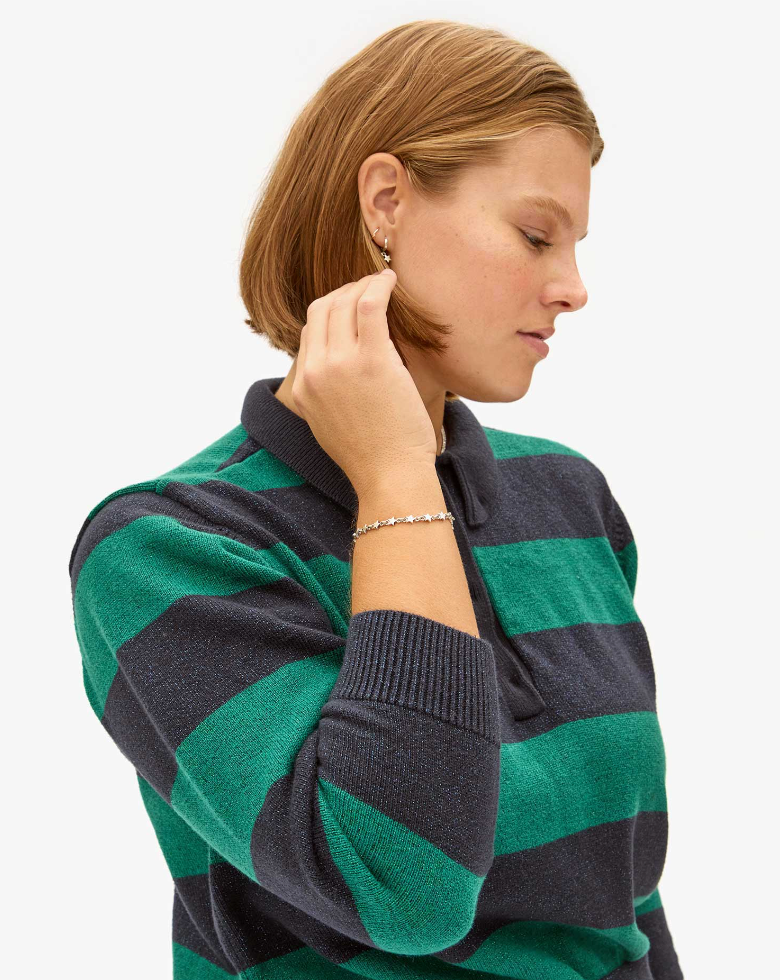 A woman with short hair wearing a green and navy striped sweater, touching her earring while looking down and to the side against a plain white background, exudes a unique Clare Vivier style with the Star Strand Bracelet Sterling Silver.