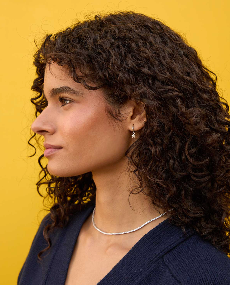 Profile view of a woman with curly hair wearing a dark blazer and Arizona-style necklace, against a yellow background. She has a small earring and is looking to her left, showcasing the Clare Vivier Star Mini Hoop Sterling Silver.