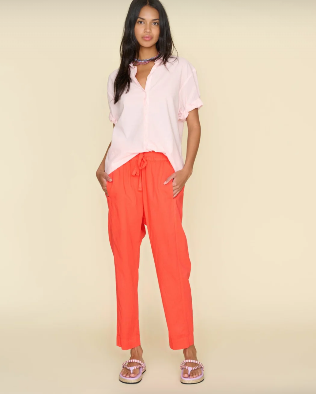 A woman with long dark hair stands against a plain beige background, reminiscent of a cozy Scottsdale bungalow. She wears a light pink shirt with rolled-up sleeves, Xirena's bright orange Punch Draper Pant, and pink sandals with decorative beading. She has her hands in her pockets and gazes at the camera.