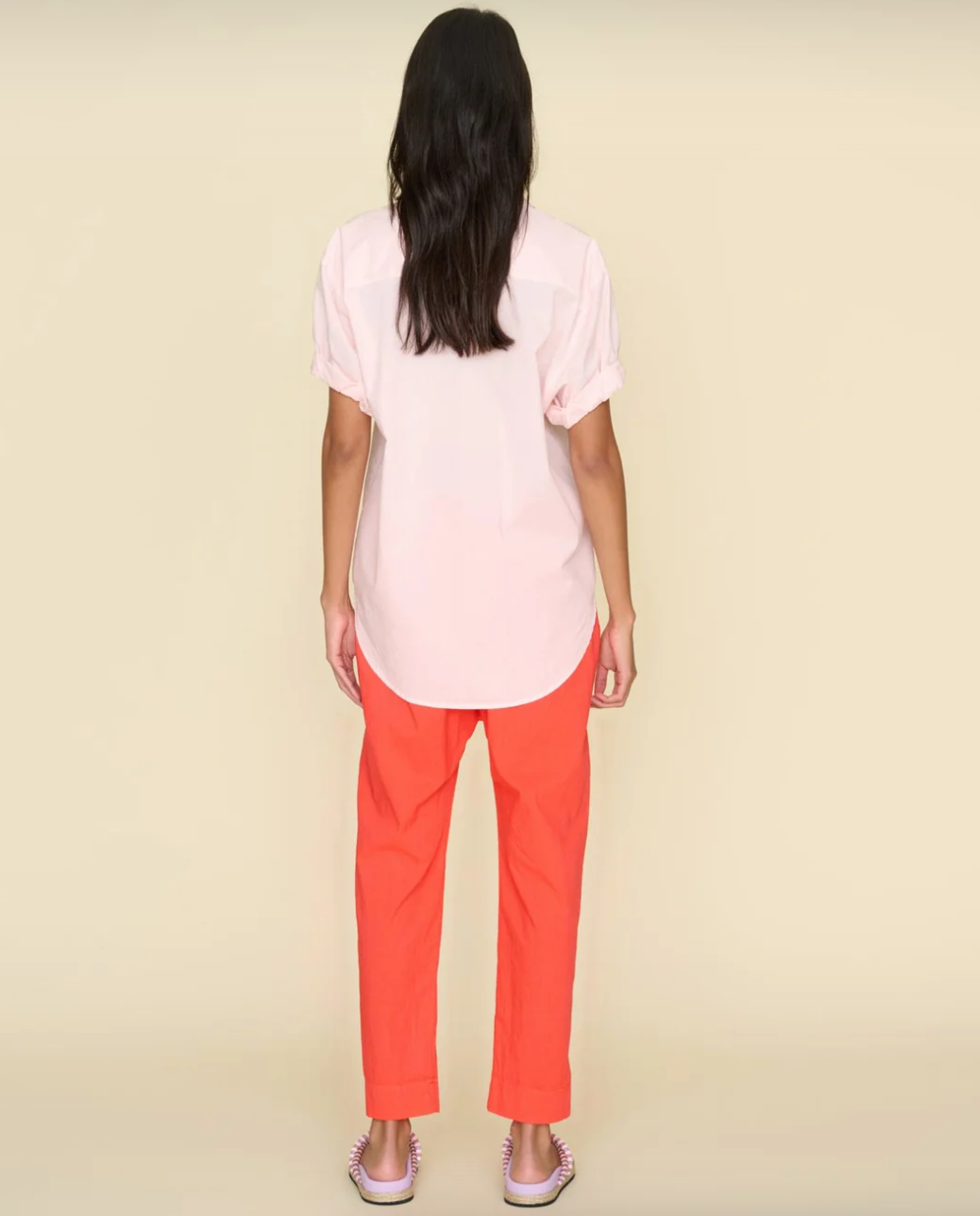 A person with long dark hair is standing with their back to the camera, set against a plain beige background. They are wearing a light pink short-sleeve shirt, bright orange Xirena Punch Draper Pant, and pink striped sandals. This could be a relaxed scene from a cozy bungalow in Scottsdale, Arizona.
