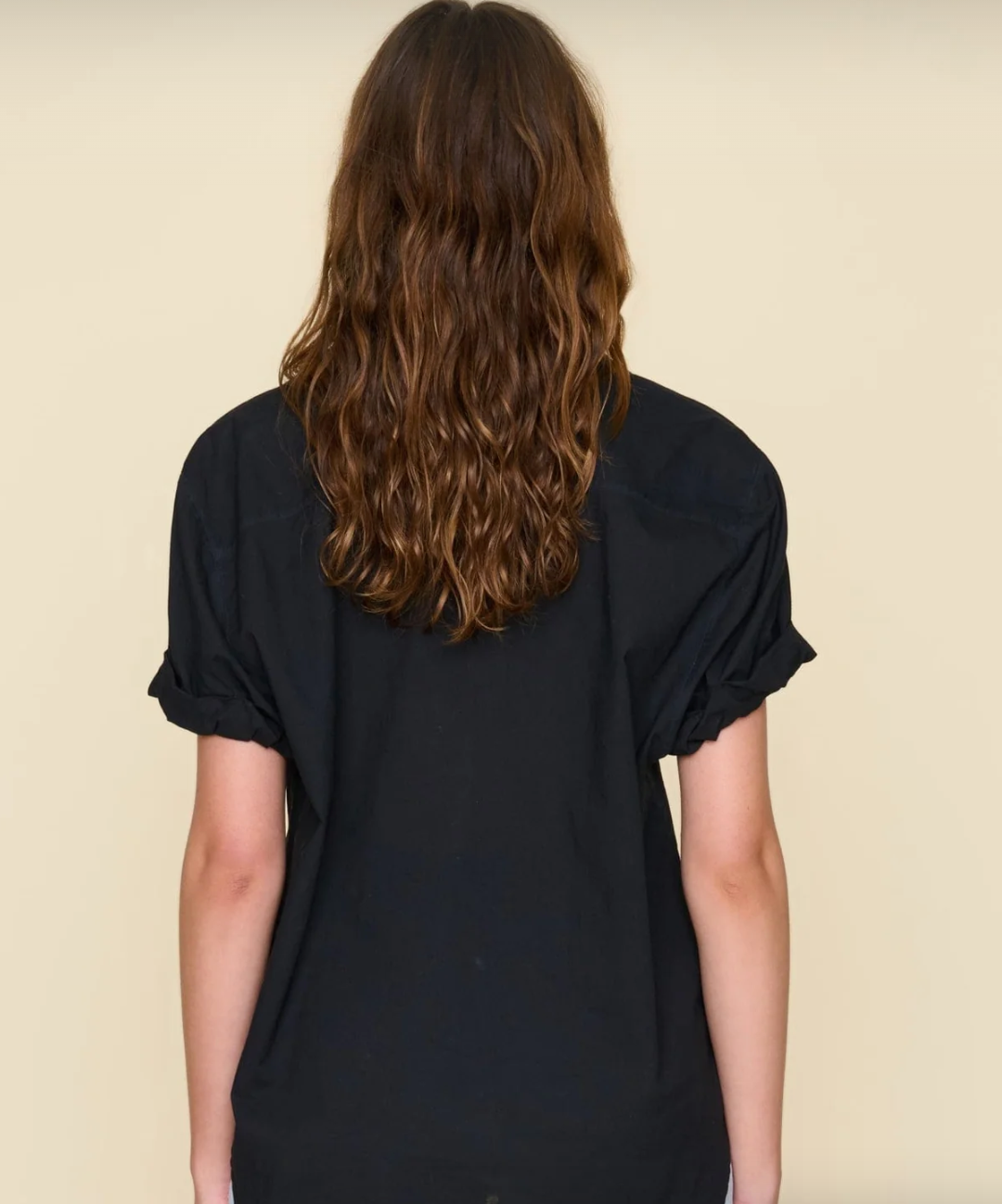 A woman with medium-length wavy brown hair, standing with her back to the camera, wearing a Xirena plain black Channing shirt against an Arizona-style beige background.