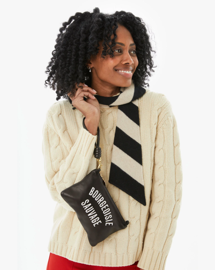 A smiling woman with curly hair sporting a cream cable-knit sweater and a black-and-white striped scarf, styled to reflect the Arizona vibe, holds a small Wallet Clutch w/ Tabs Black Bourgeoisie Sauvage from Clare Vivier that reads "Bourgeoisie Scum.