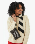 A smiling woman with curly hair sporting a cream cable-knit sweater and a black-and-white striped scarf, styled to reflect the Arizona vibe, holds a small Wallet Clutch w/ Tabs Black Bourgeoisie Sauvage from Clare Vivier that reads "Bourgeoisie Scum.