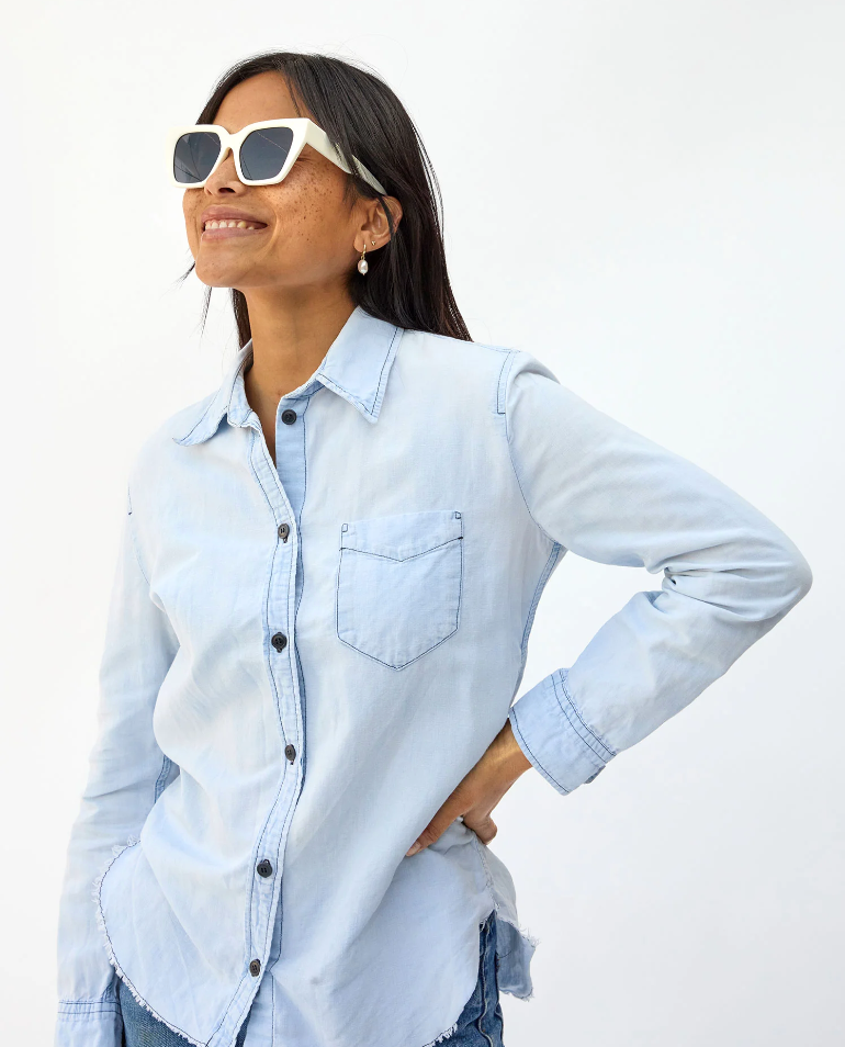A woman in Clare V. Heather Sunglasses Cream by Clare Vivier and an Arizona style light blue denim shirt smiling and looking left, against a plain white background. Her hand is placed on her hip.