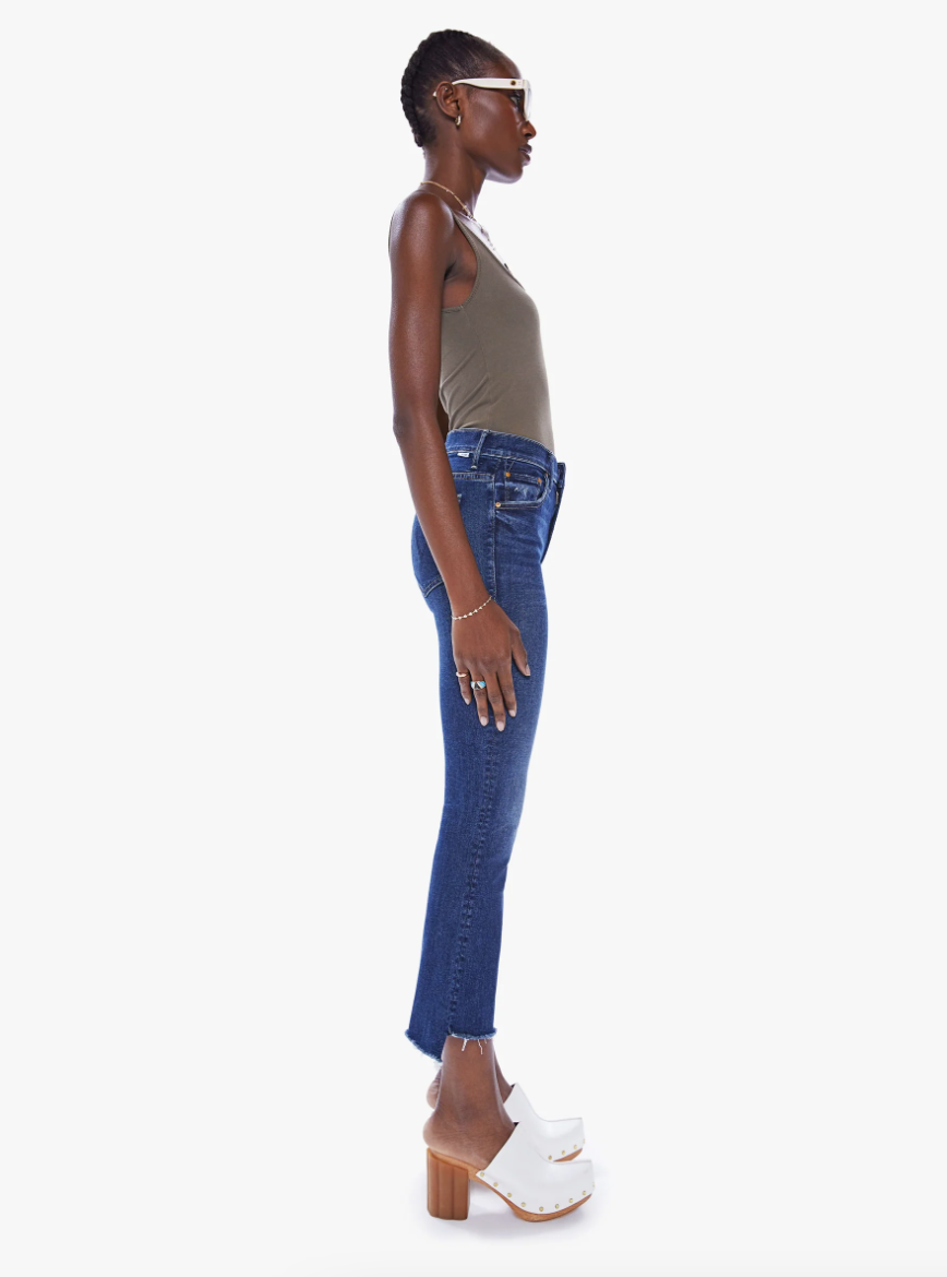 A profile view of a Black woman standing, wearing the Teaming Up Insider Crop Step Fray Wash high-waisted bootcut jeans, and white platform sandals. She is looking straight ahead and has her hair pulled back.