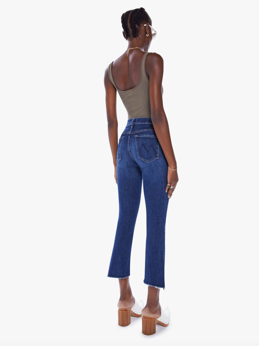 A woman seen from behind, wearing a sleeveless olive green top and high-waisted bootcut blue jeans with a step frayed hem, standing against a white background. She is paired with white heels in The Insider Crop Step Fray Wash: Teaming Up by [Brand Name].