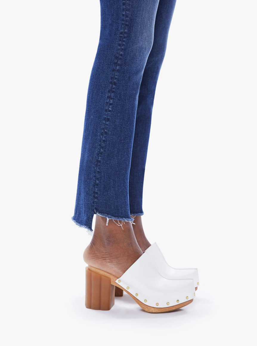 A person wearing The Insider Crop Step Fray Wash: Teaming Up blue jeans with a step frayed hem and one white clog with a wooden heel, adorned with golden studs, standing against a white background.