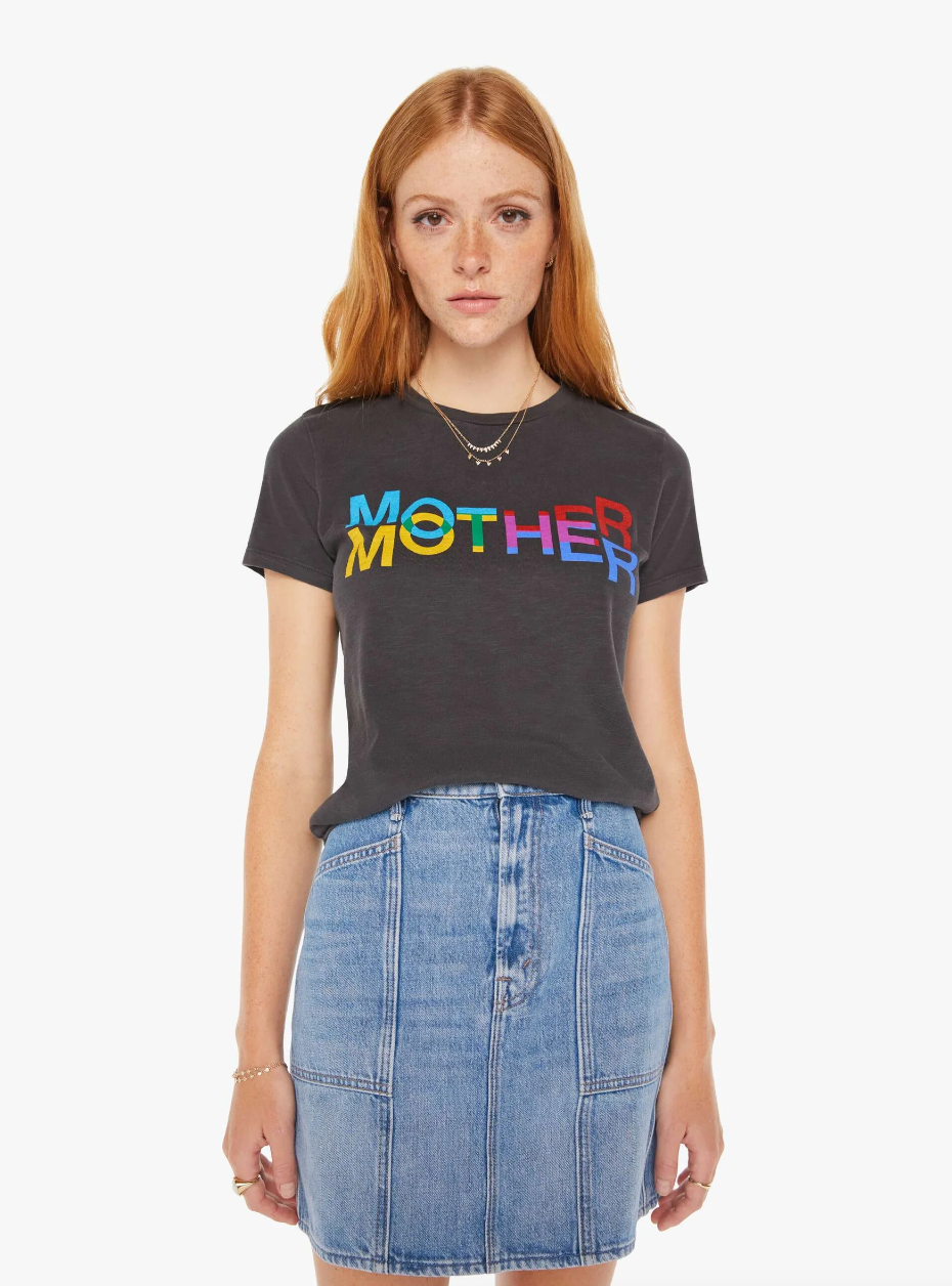 A woman with long red hair and freckles is wearing a black T-shirt with the words "The Lil Sinful Mother Kaleidoscope" printed in colorful letters. She is also wearing a light denim skirt and layered necklaces, standing against a plain white background reminiscent of the sunny bungalows in Scottsdale, Arizona.