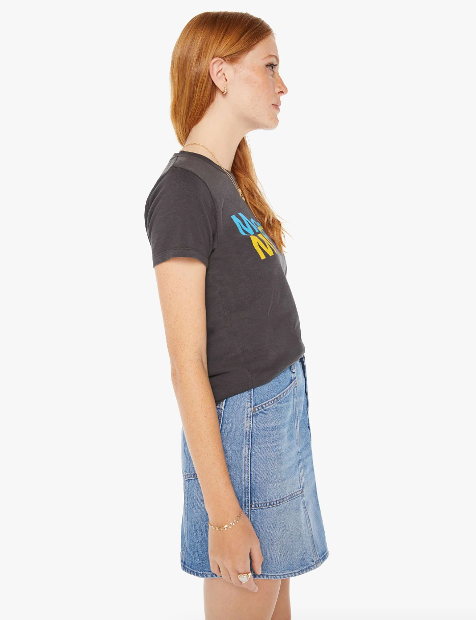A person with long red hair stands in profile wearing The Lil Sinful Mother Kaleidoscope dark gray t-shirt with yellow and blue text, paired with a blue denim skirt. They are accessorized with a bracelet. The background is plain and white, reminiscent of the minimalist decor often seen in cozy Scottsdale bungalows.
