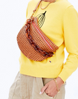 A person wearing a yellow sweater with a black fruit outline and beige pants is carrying a woven brown handbag that has the Clare Vivier Shortie Strap with brass spring links over their shoulder. They are accessorized with a gold bracelet and have their hand in their pocket.