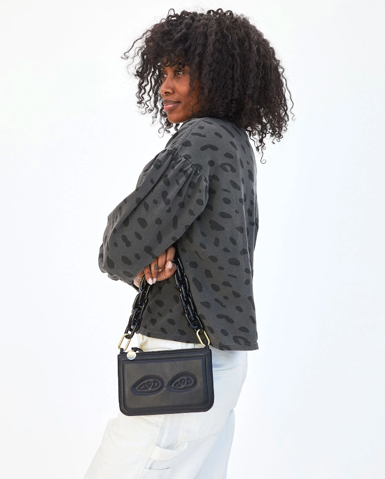 A person with curly hair is turned slightly to the side, showcasing a Clare Vivier Shortie Strap. They are wearing a grey, spotted long-sleeve top and light-colored pants. The background is plain and white.