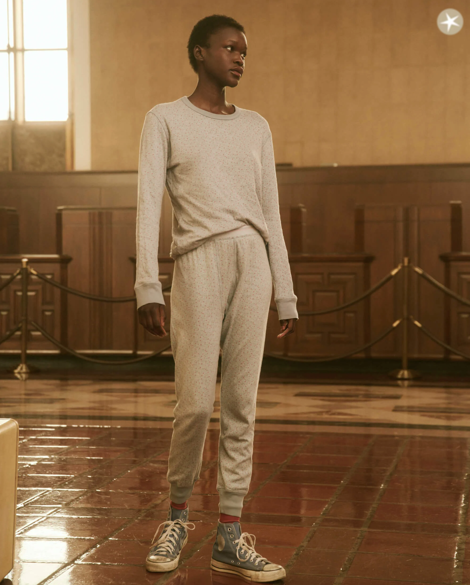 A person in a gray sweatshirt and sweatpants stands in a large room with wooden walls and a tiled floor, reminiscent of an old-fashioned hall or waiting area. Casually dressed in THE SURPLUS CREW BLUEBELL GREENHOUSE FLORAL by The Great Inc., they could easily be mistaken for someone lounging in a cozy bungalow rather than this roped-off space.