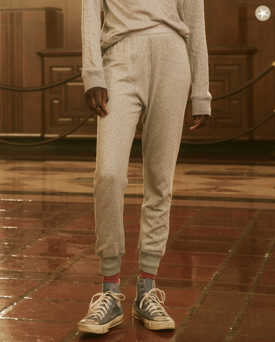 Person in a light gray long-sleeve shirt and matching sweatpants with tiny dot patterns, standing indoors on a polished floor of their cozy Scottsdale bungalow. They are wearing worn gray high-top sneakers with blue accents and red socks from The Great Inc.’s THE LONG JOHN BLUEBELL GREENHOUSE FLORAL collection.