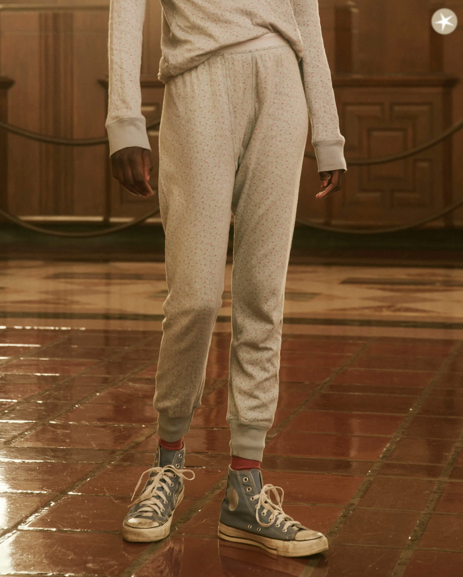 A person stands on a polished floor in a cozy bungalow, wearing light gray joggers with a subtle dotted pattern, a matching sweatshirt, and THE LONG JOHN BLUEBELL GREENHOUSE FLORAL high-top sneakers with white laces by The Great Inc. The background features wooden paneling and architectural details reminiscent of Scottsdale, Arizona style.