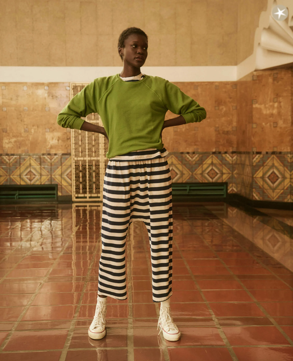 A person stands confidently in an indoor setting with hands on hips. They are wearing a green long-sleeve shirt, THE JERSEY CROP NAVY AND CREAM SCHOLAR STRIPE by The Great Inc., and white high-top sneakers. The background features a tiled wall and floor with an art deco style, reminiscent of a chic Scottsdale Arizona bungalow.