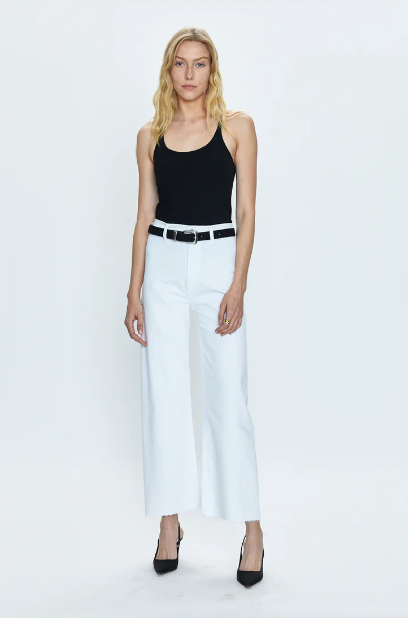 A woman stands against a plain background, wearing a Pistola Los Angeles-based style black tank top, white high-waisted pants with a black belt, and black high heels.