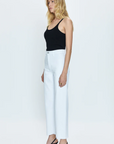 A Los Angeles-based woman stands against a white background, wearing a black tank top paired with white high-waisted PENNY HIGH RISE WIDE LEG CROP - BLIZZARD trousers from Pistola and black heels.