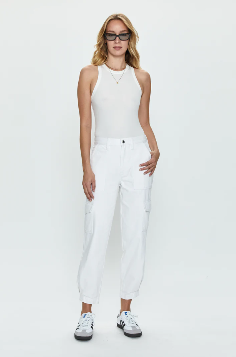 A Los Angeles-based woman in a white sleeveless top and Pistola&#39;s Josephine High Rise Tapered Cargo - White stands confidently against a plain white background, wearing white sneakers and stylish sunglasses.