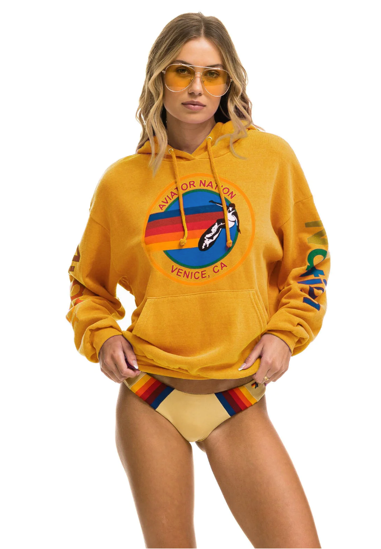 A woman wearing an Aviator Nation relaxed pullover hoodie in gold and yellow sunglasses stands confidently, pairing her hoodie with a beige bikini bottom in true Arizona style. The hoodie features a colorful graphic and text.