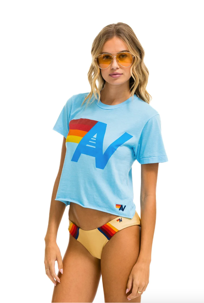 A person with long blonde hair is wearing orange-tinted sunglasses, an Aviator Nation LOGO BOYFRIEND TEE - SKY with a rainbow-colored 'A' graphic, and yellow bikini bottoms with rainbow stripes. Posing in front of a white backdrop, they look ready for summer vibes reminiscent of a chic bungalow in Scottsdale, Arizona.