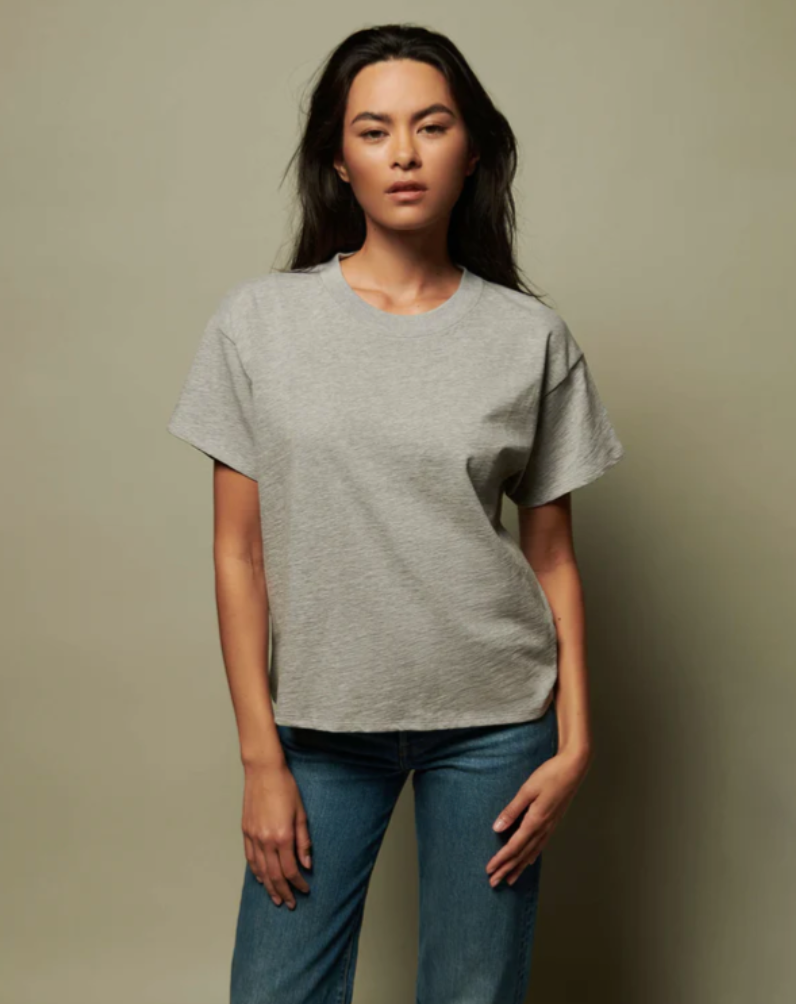 A person with long dark hair wearing a JESSA TOP HEATHER GREY by Nation LTD and blue jeans stands against a plain beige background. They have a neutral expression, with their left hand resting on their right hip while their right arm hangs by their side. It feels as if they're casually posing outside a charming bungalow in Scottsdale, Arizona.