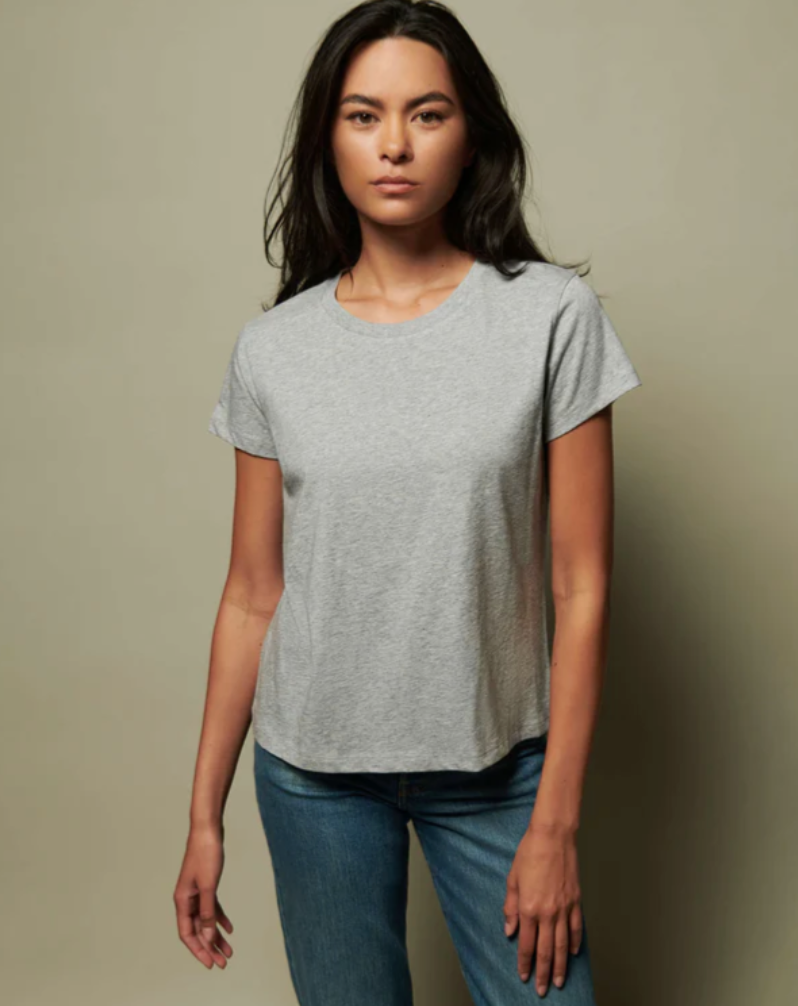 A person with long dark hair wearing a GOLDIE TEE HEATHER GREY by Nation LTD and blue jeans stands against a neutral background, reminiscent of a cozy bungalow in Scottsdale, Arizona, staring directly at the camera.