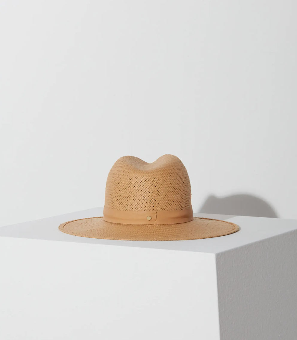A light brown straw fedora hat, Simone Hat Sand-style from Janessa Leone, resting on a white pedestal against a white background, casting a soft shadow on the surface.