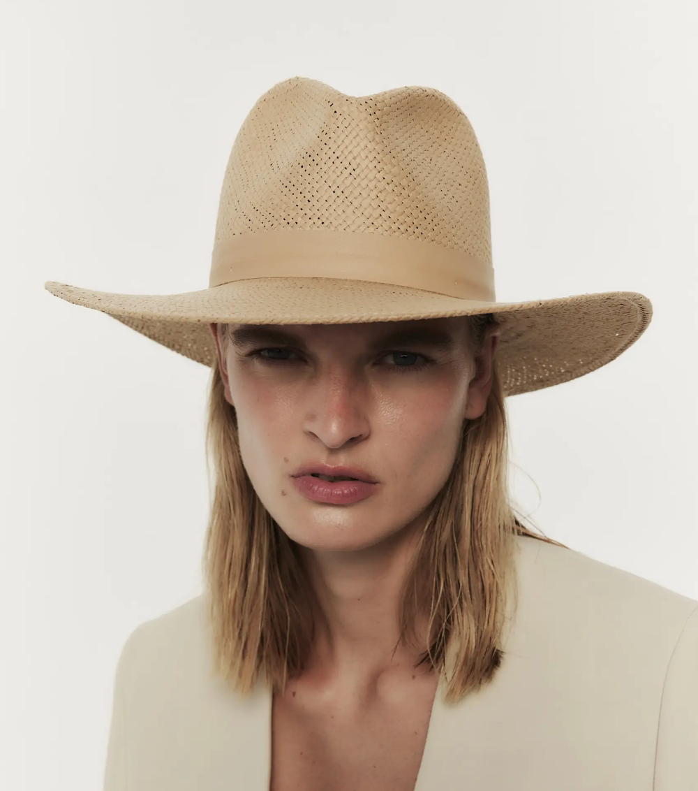 A person with shoulder-length blond hair wearing a light beige, wide-brimmed Janessa Leone SIMONE HAT SAND, looking intently towards the viewer against a white background.
