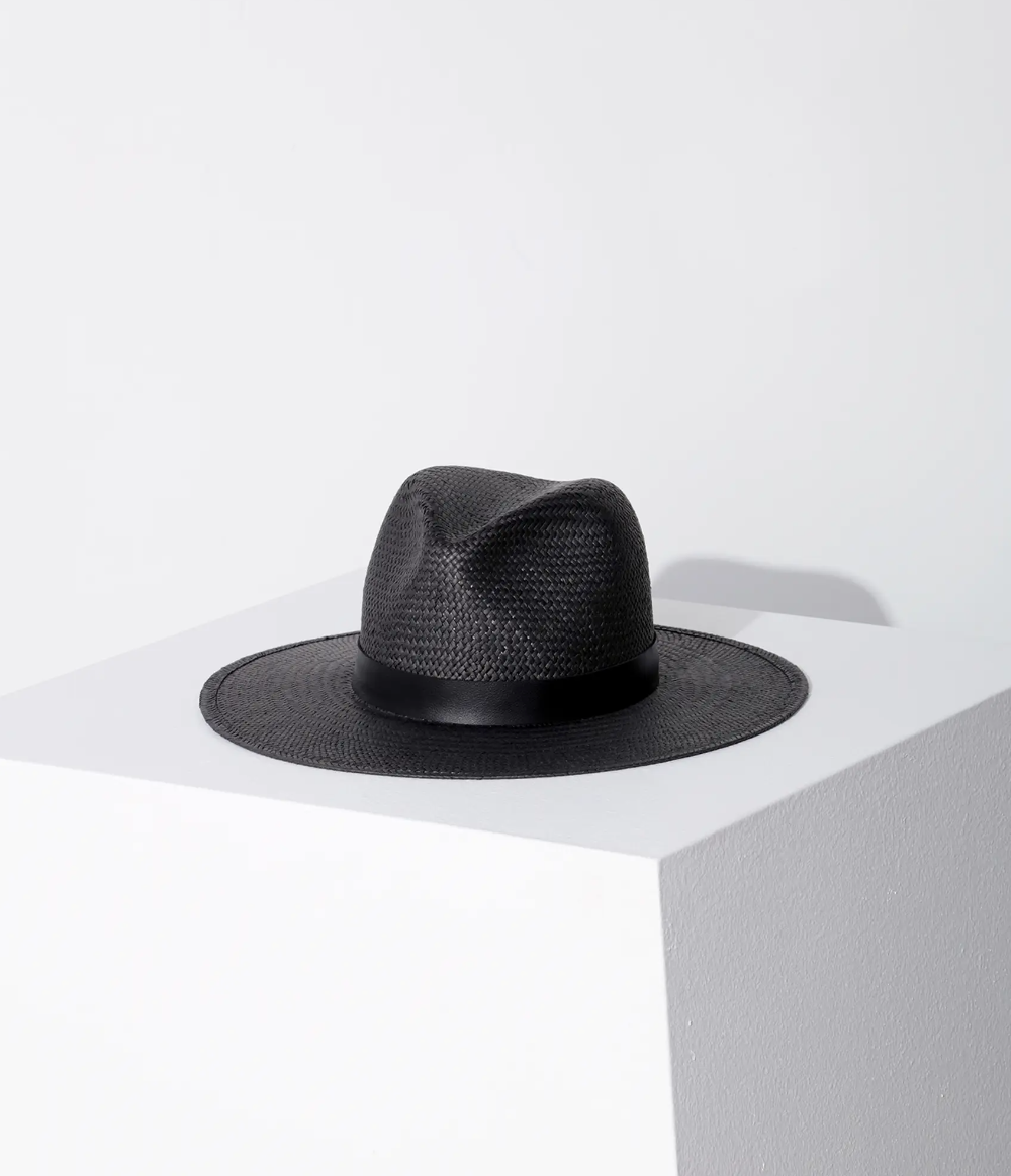 A black Simone hat by Janessa Leone with Arizona style placed on a white corner shelf against a plain light gray background, highlighting its texture and shape under soft lighting.