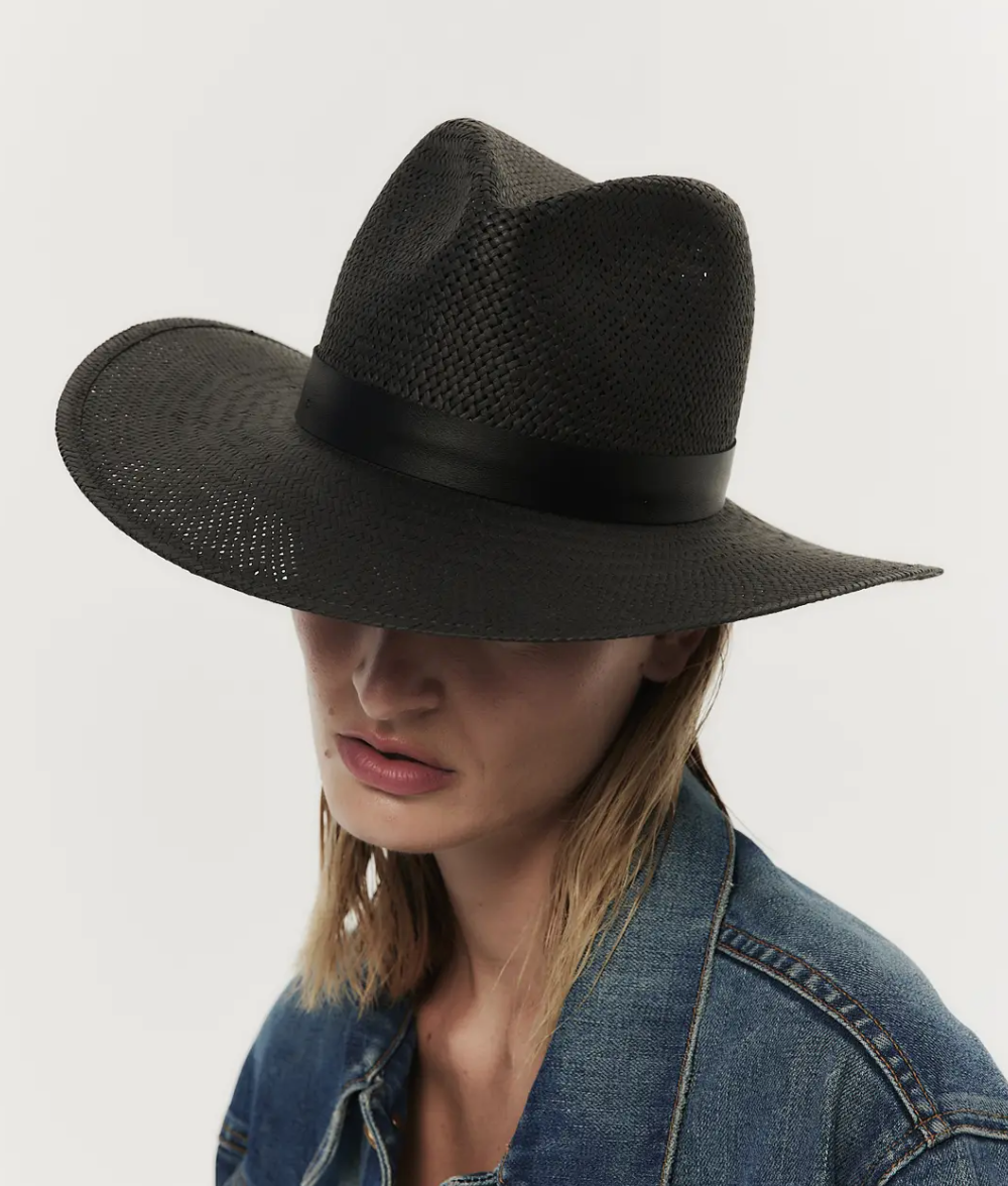 A woman in a denim jacket wears a large SIMONE HAT BLACK by Janessa Leone that casts a shadow over her face, emphasizing the stylish texture of the hat.