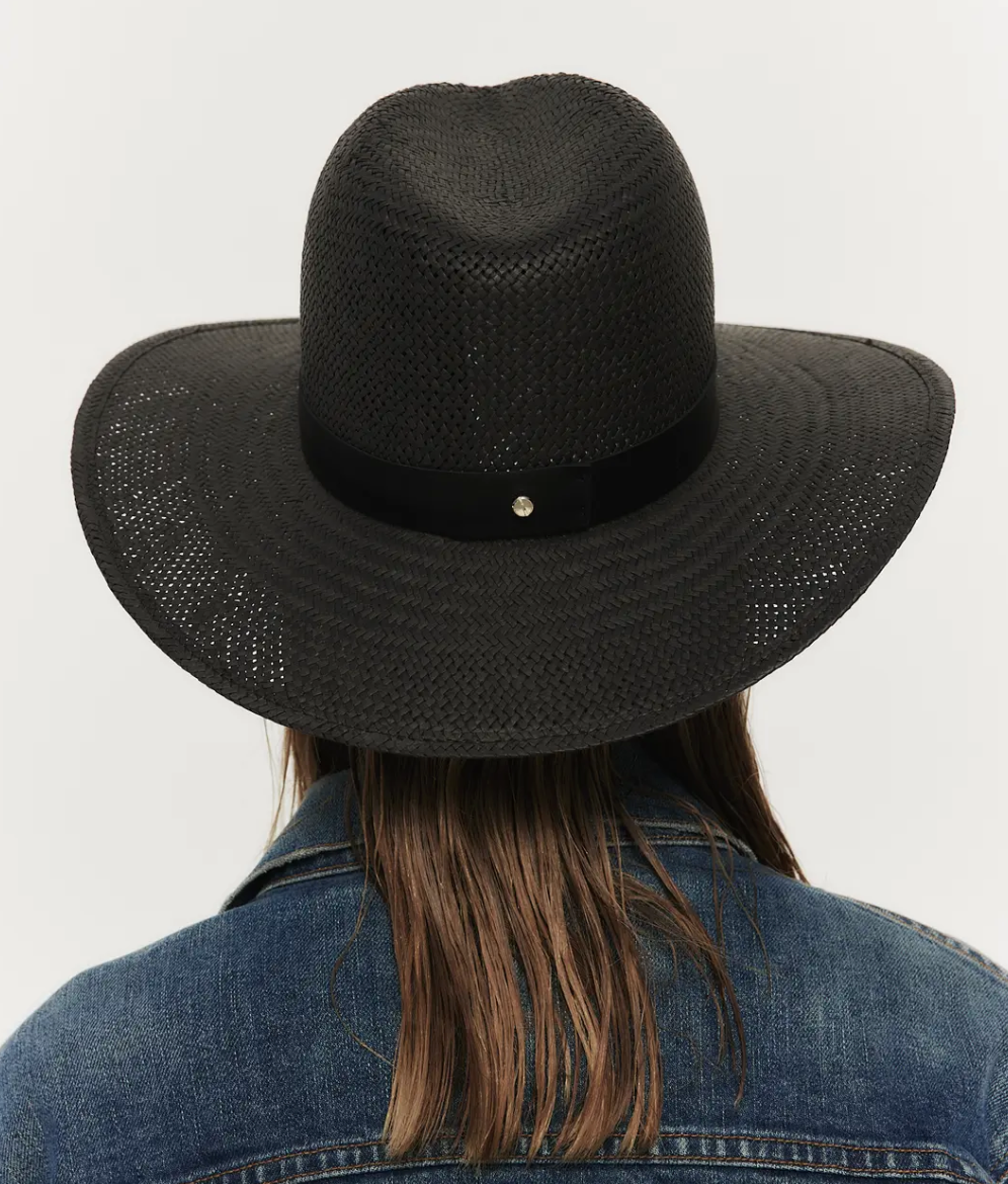 Rear view of a person sporting an Janessa Leone SIMONE HAT BLACK wide-brimmed hat and a denim jacket, focusing on the hat which has a perforated pattern and a black ribbon around the crown.
