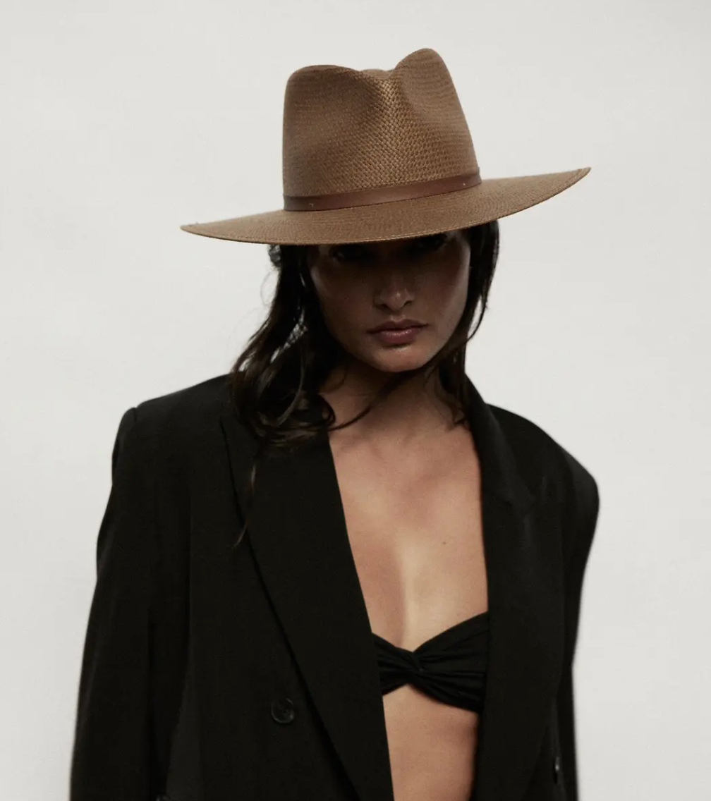 A person wearing a stylish Janessa Leone brown Sherman hat and a black blazer over a black bikini top, looking intensely at the camera with a shadowed face, evoking the effortless style of Arizona.