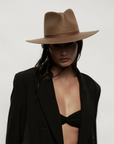 A person wearing a stylish Janessa Leone brown Sherman hat and a black blazer over a black bikini top, looking intensely at the camera with a shadowed face, evoking the effortless style of Arizona.
